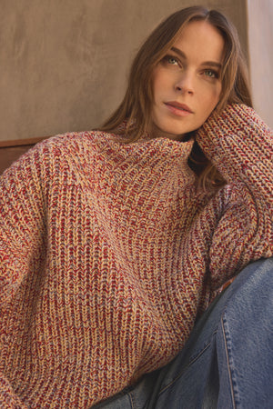 A woman with long brown hair wears the DENNIE SWEATER by Velvet by Graham & Spencer—a chunky wool blend sweater with a marled texture—paired with blue jeans. She sits with her head resting on her hand, looking slightly upward.