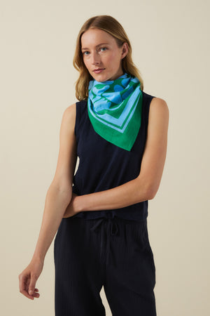 A woman stands against a plain light background, wearing a navy sleeveless top and pants, accessorized with the Velvet by Graham & Spencer SOLI SCARF—a printed cotton square scarf—tied stylishly around her neck.