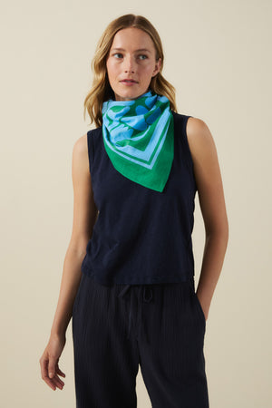 A woman stands against a beige background wearing a sleeveless black top, black pants, and the Velvet by Graham & Spencer SOLI SCARF—a versatile printed cotton accessory—styled around her neck.