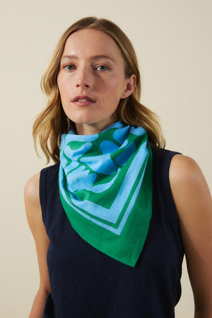 A woman stands against a beige backdrop wearing the Velvet by Graham & Spencer SOLI SCARF—a blue and green printed cotton square scarf—styled around her neck to showcase its versatile design.