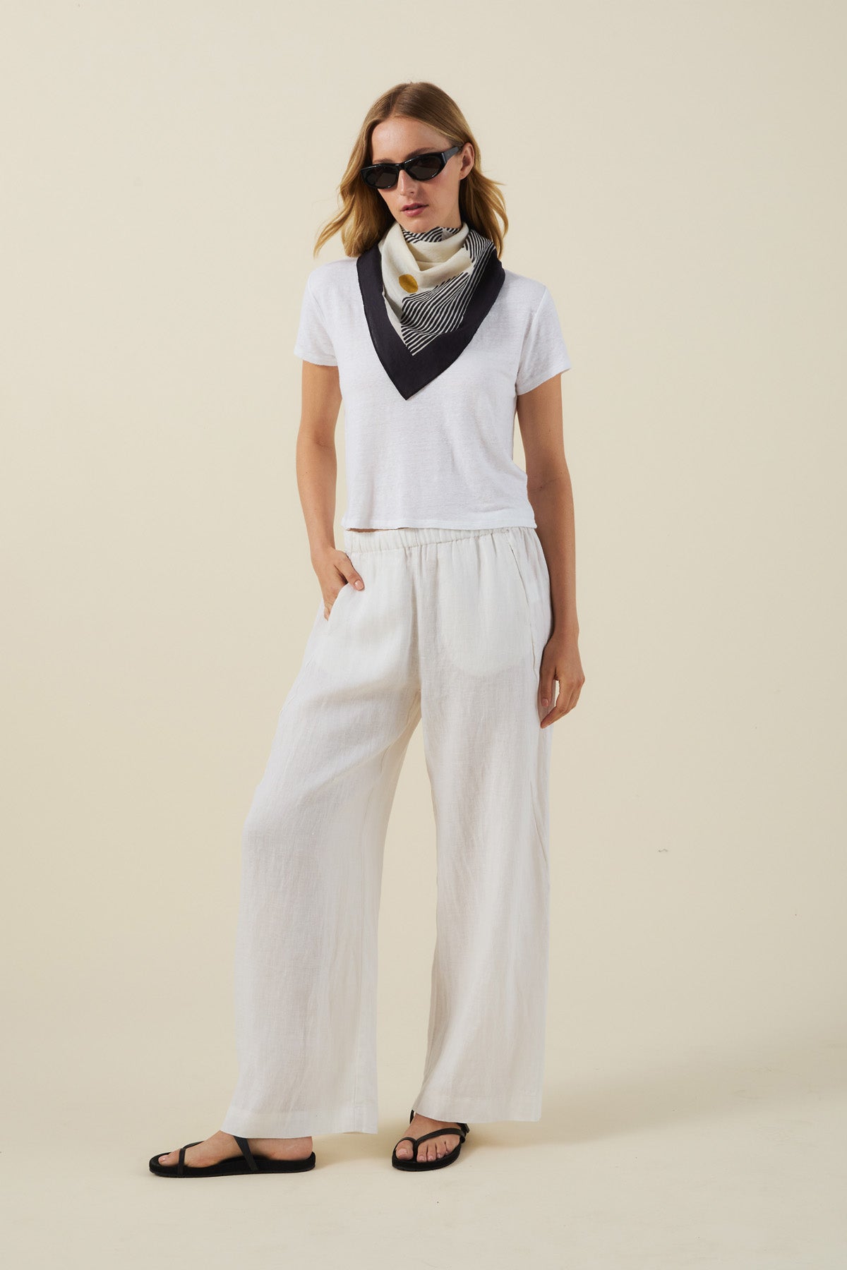 A person wearing a white t-shirt, white wide-leg pants, black sandals, sunglasses, and the Velvet by Graham & Spencer SOLI SCARF stands against a plain light background, highlighting the effortless style of this versatile accessory.-41527506108609