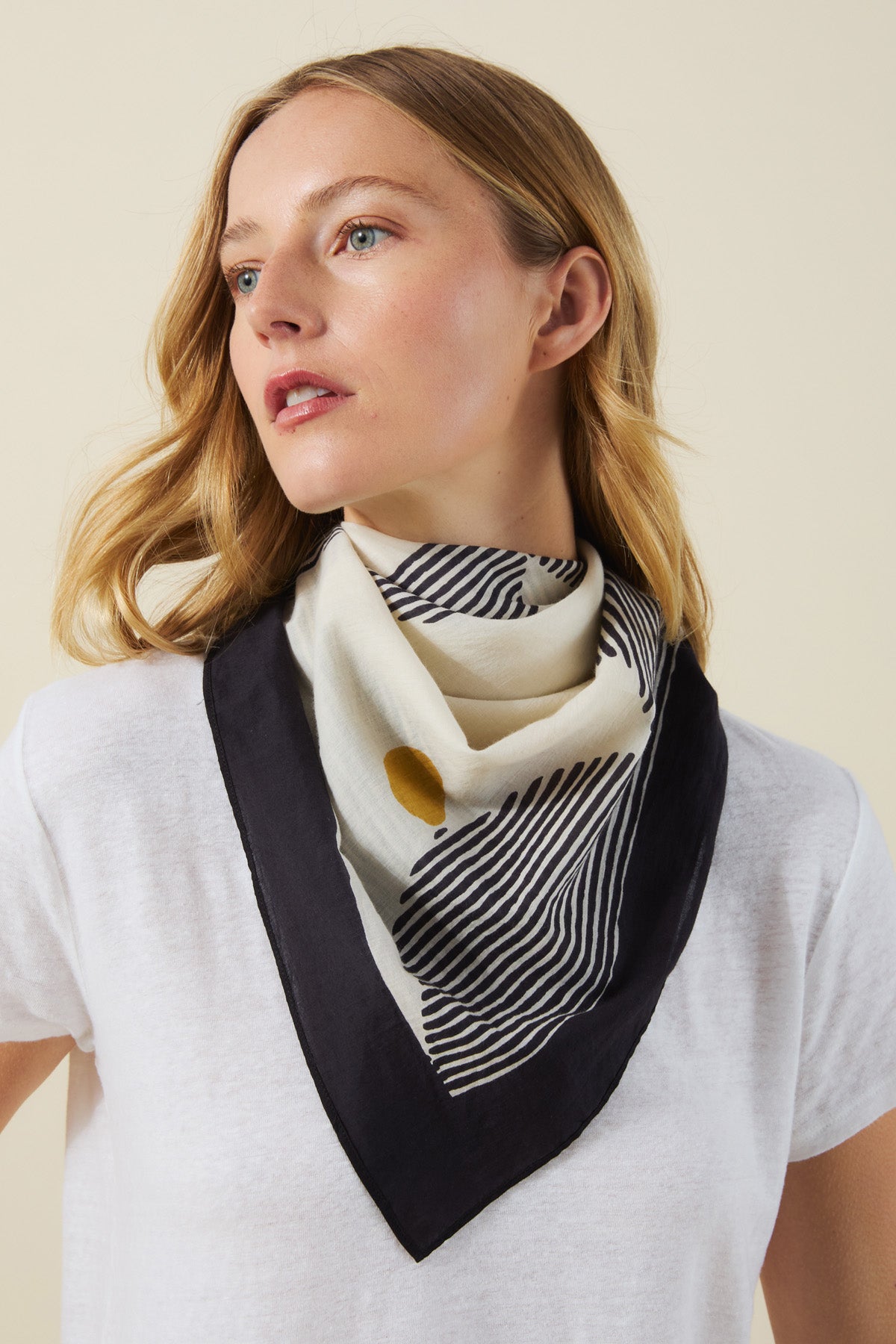 A woman wearing a white T-shirt and the Velvet by Graham & Spencer SOLI SCARF, a square black and white printed cotton scarf with a yellow dot, looking slightly to the side.
