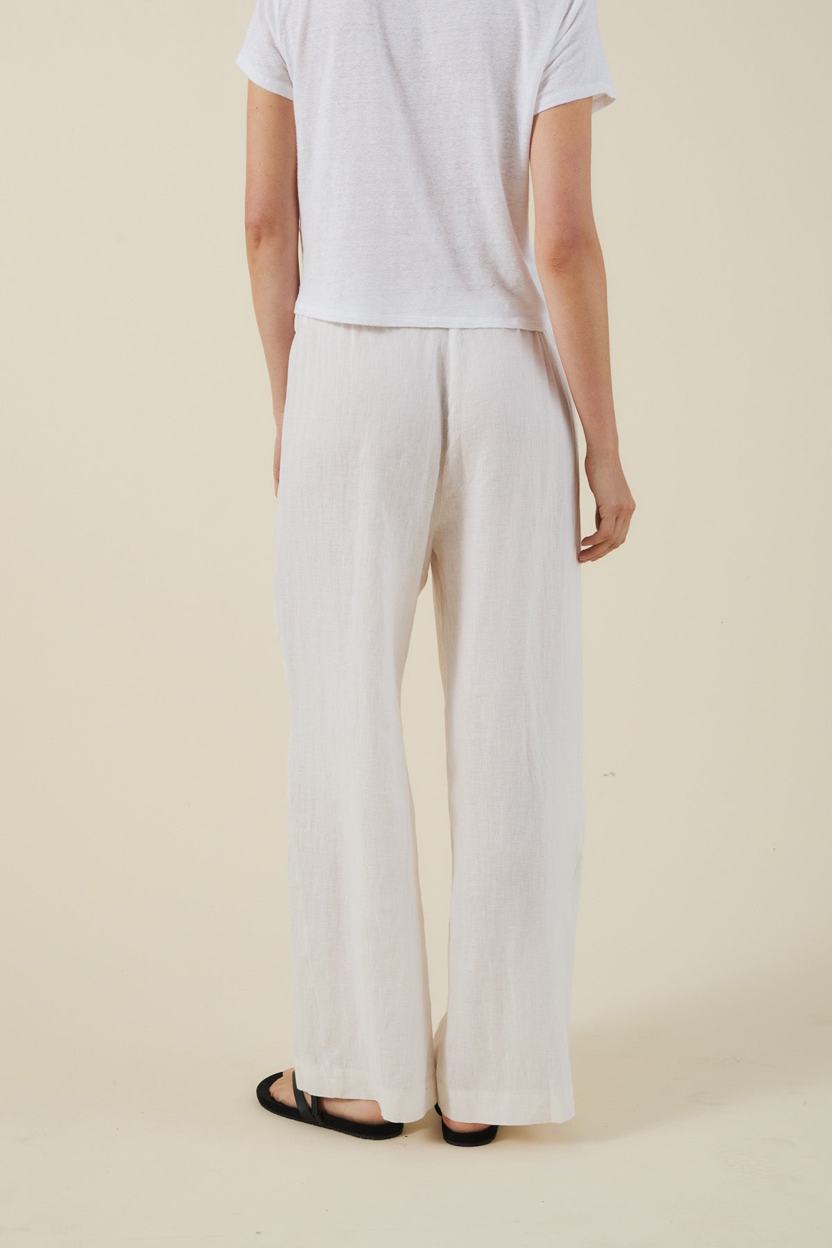 A person is seen from behind, wearing the Velvet by Graham & Spencer STEPHANIE LINEN PANT with a white short-sleeve shirt and black sandals, standing against a plain background.
