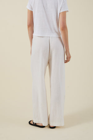 A person is seen from behind, wearing the Velvet by Graham & Spencer STEPHANIE LINEN PANT with a white short-sleeve shirt and black sandals, standing against a plain background.