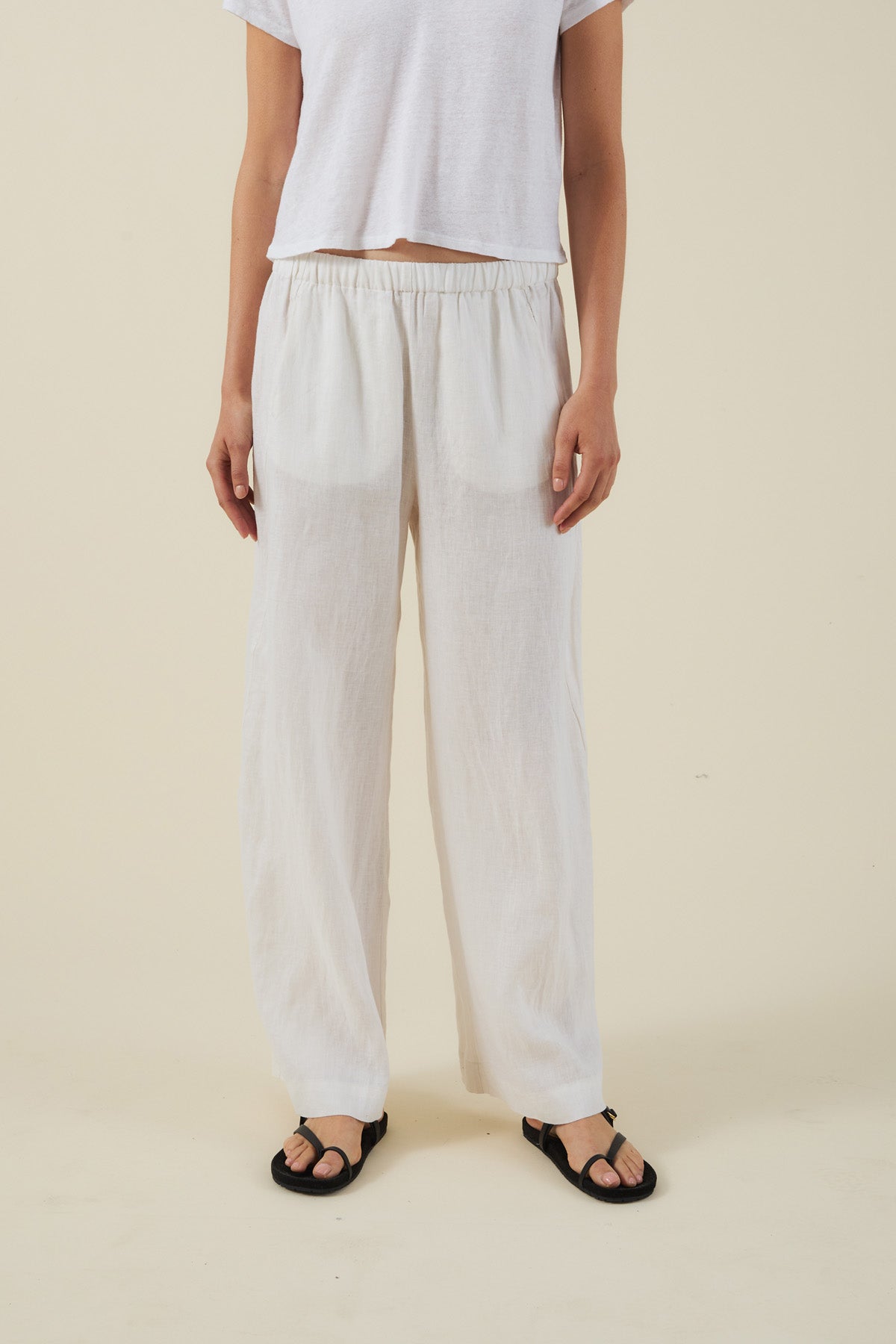 A person wears a white short-sleeve shirt, Velvet by Graham & Spencer's STEPHANIE LINEN PANT with an elastic waist, and black sandals, standing against a plain beige background.-41645109805249