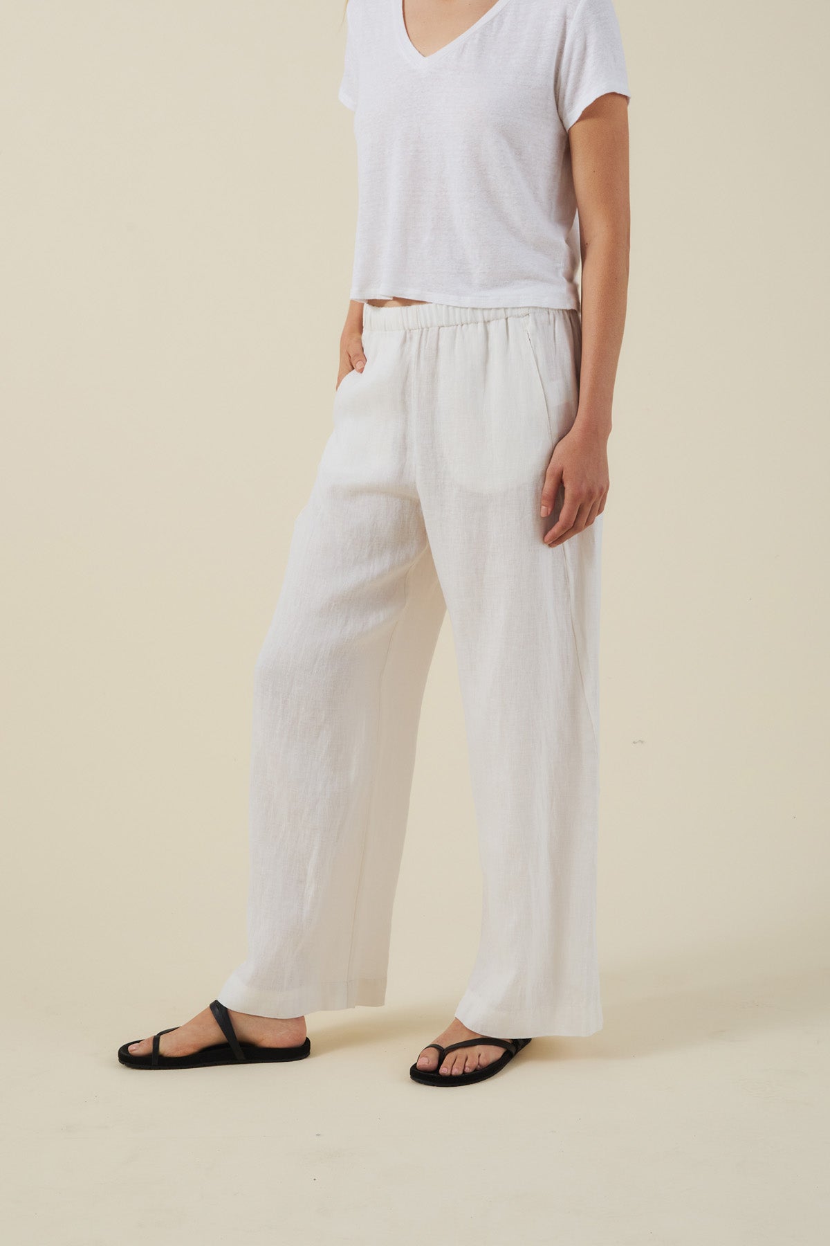 A person stands against a beige background, wearing a loose white short-sleeve top, black sandals, and the STEPHANIE LINEN PANT by Velvet by Graham & Spencer—wide-leg linen pants with an elastic waist.-41645109936321