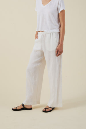 A person stands against a beige background, wearing a loose white short-sleeve top, black sandals, and the STEPHANIE LINEN PANT by Velvet by Graham & Spencer—wide-leg linen pants with an elastic waist.