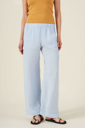 Person wearing a sleeveless mustard top, black sandals, and the STEPHANIE LINEN PANT by Velvet by Graham & Spencer in light blue with an elastic waist, standing against a neutral background.