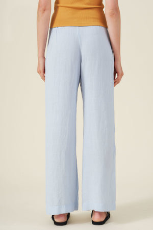 Person facing away wears Velvet by Graham & Spencer's STEPHANIE LINEN PANT in light blue with an elastic waist, paired with a mustard sleeveless top and black shoes against a neutral background.