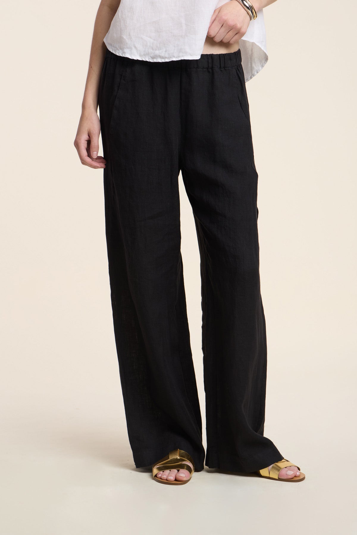 Model wearing the STEPHANIE LINEN PANT by Velvet by Graham & Spencer, a relaxed fit black wide-leg pant, paired with a white top and gold sandals, standing against a plain light background.-41372219211969