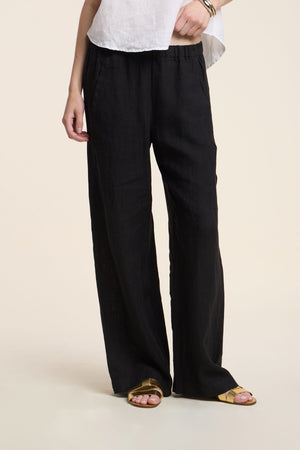 Model wearing the STEPHANIE LINEN PANT by Velvet by Graham & Spencer, a relaxed fit black wide-leg pant, paired with a white top and gold sandals, standing against a plain light background.