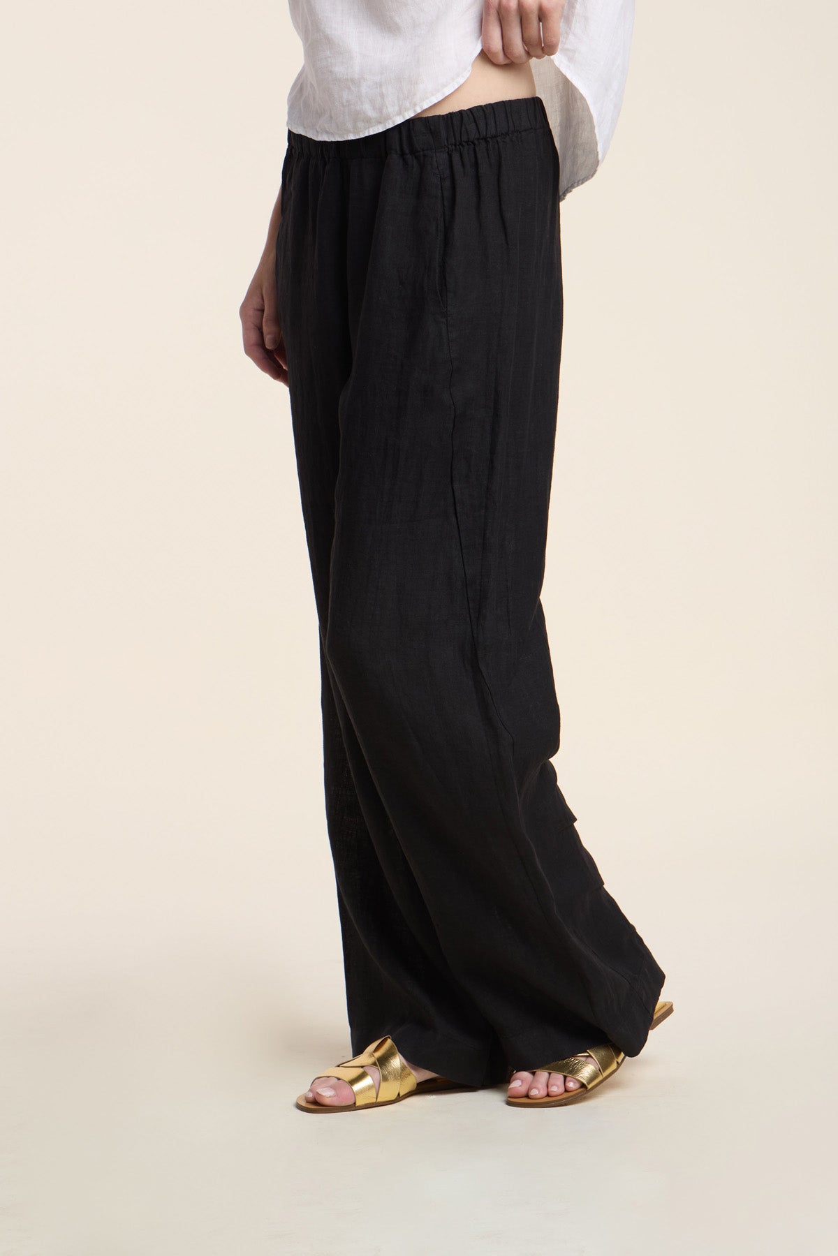 A person models the STEPHANIE LINEN PANT by Velvet by Graham & Spencer—relaxed fit, wide-leg black pants—paired with a white top and gold sandals, shown from the waist down against a plain light background.-41372219244737