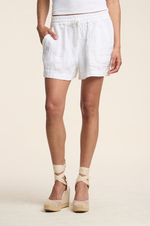 A person in a white tank top, Velvet by Graham & Spencer SUTTON HEAVY LINEN SHORTS with drawstring waist and pockets, and beige espadrille wedges stands against a plain background.