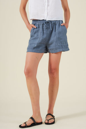 A person in a white short-sleeve button-up shirt, Velvet by Graham & Spencer SUTTON HEAVY LINEN SHORTS in blue with a drawstring waist and front pockets, and black sandals stands against a plain background.