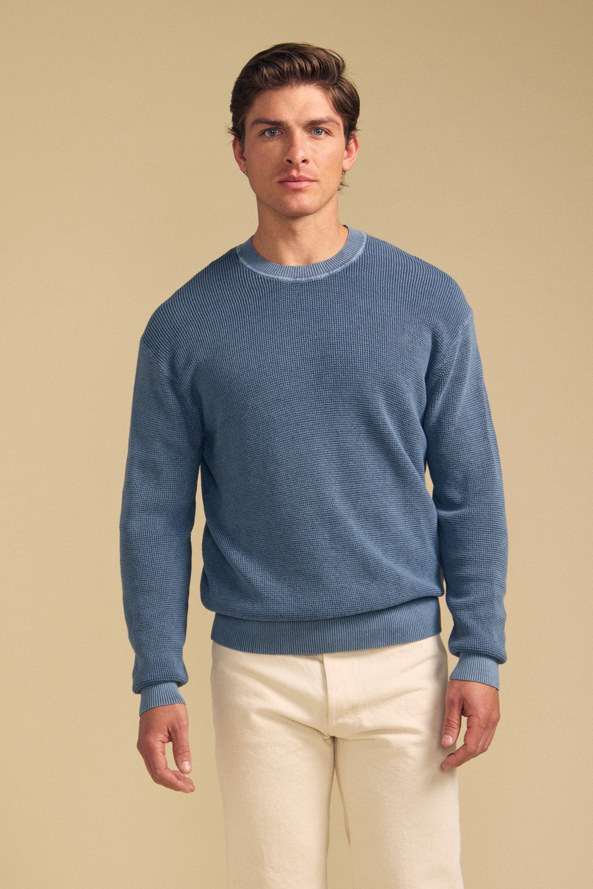 A man stands facing forward, wearing the PHINN SWEATER by Velvet by Graham & Spencer—a blue crew neck sweater—paired with cream-colored pants against a plain beige background.