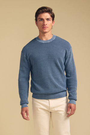 A man stands facing forward, wearing the PHINN SWEATER by Velvet by Graham & Spencer—a blue crew neck sweater—paired with cream-colored pants against a plain beige background.