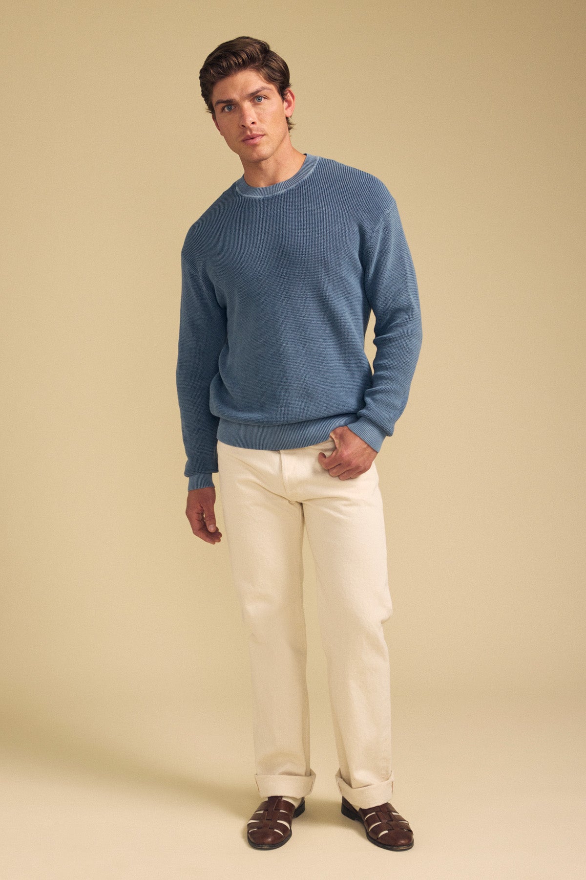 A man stands against a beige background wearing the PHINN SWEATER by Velvet by Graham & Spencer in blue, paired with cream pants and brown shoes. He has a neutral facial expression and a relaxed posture.