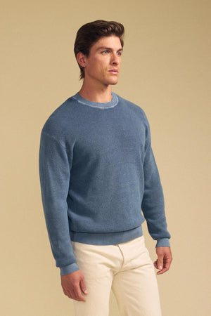 A person stands against a plain beige background, wearing the Velvet by Graham & Spencer PHINN SWEATER in light blue and off-white pants.