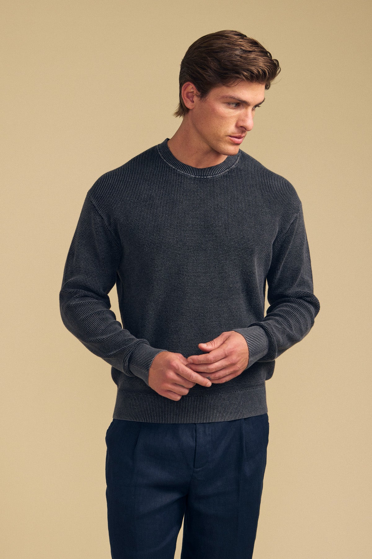 A man stands against a beige background, wearing the PHINN SWEATER by Velvet by Graham & Spencer in dark gray ribbed knit and navy trousers, looking slightly to his right with his hands loosely clasped.