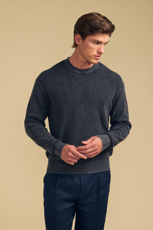 A man stands against a beige background, wearing the PHINN SWEATER by Velvet by Graham & Spencer in dark gray ribbed knit and navy trousers, looking slightly to his right with his hands loosely clasped.