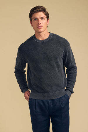 A man wearing the PHINN SWEATER by Velvet by Graham & Spencer in dark gray, paired with dark pants, stands against a beige background looking at the camera with his hands in his pockets.