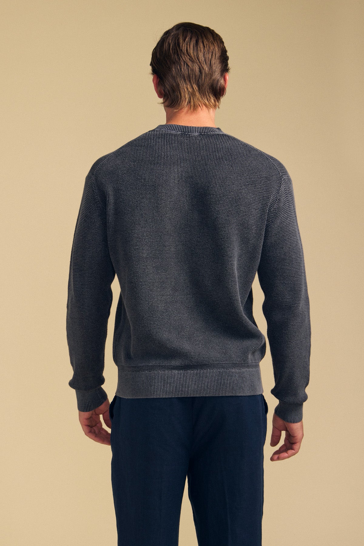 A man stands with his back to the camera, wearing the PHINN SWEATER in dark gray from Velvet by Graham & Spencer and navy blue pants against a plain beige background.-41759810191553