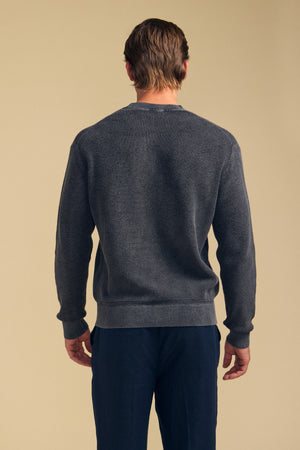 A man stands with his back to the camera, wearing the PHINN SWEATER in dark gray from Velvet by Graham & Spencer and navy blue pants against a plain beige background.