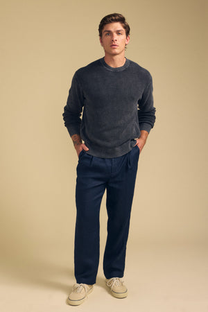 A man stands facing forward, wearing the PHINN SWEATER by Velvet by Graham & Spencer, navy pants, and cream-colored sneakers against a beige background.