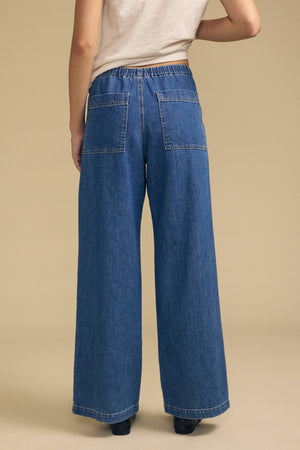 A person wearing the SYD JEAN by Velvet by Graham & Spencer—a relaxed fit, wide-leg denim jean featuring large back pockets and an elastic drawstring waist—paired with a beige top, stands on a neutral background, viewed from behind.