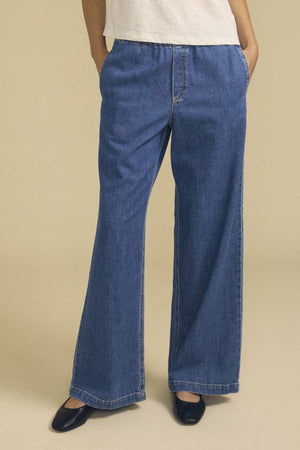 Wearing the SYD JEAN by Velvet by Graham & Spencer—relaxed fit blue jeans featuring an elastic drawstring waist—styled with a white shirt and black shoes, this person stands with hands in pockets against a plain background.