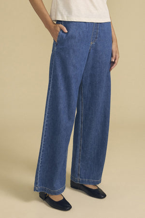 A person stands with hands in pockets against a plain beige background, wearing the Velvet by Graham & Spencer SYD JEAN—relaxed fit wide-leg denim jeans with an elastic drawstring waist—paired with a light short-sleeve shirt and black flat shoes.