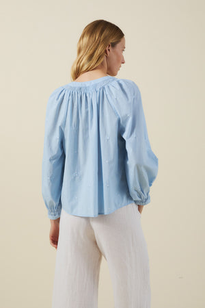 A person with blonde hair, seen from behind, wears the SYLVIE TOP by Velvet by Graham & Spencer—a light blue cotton poplin peasant blouse with subtle embroidery—paired with white loose-fitting pants against a plain beige background.
