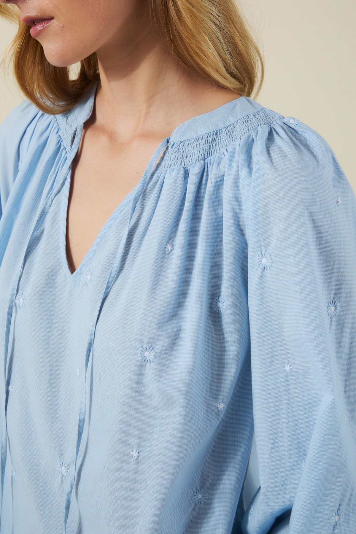 A woman wears the SYLVIE TOP by Velvet by Graham & Spencer, featuring a light blue V-neck, smocked collar, and subtle floral embroidery on soft cotton poplin.-41527517642945