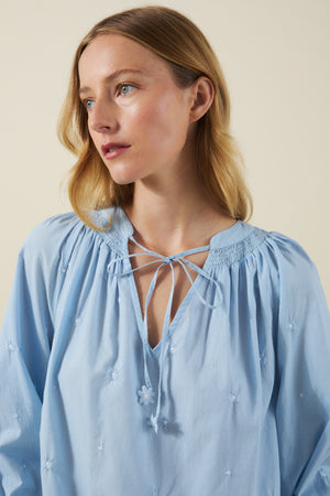A woman with blonde hair wears the SYLVIE TOP by Velvet by Graham & Spencer—a light blue cotton poplin peasant blouse featuring small floral embroidery and a tie neckline—while looking slightly to the side against a plain background.