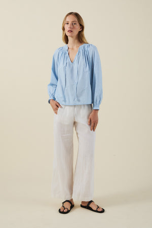 A person stands against a plain background wearing the SYLVIE TOP from Velvet by Graham & Spencer in light blue cotton poplin, paired with white wide-leg pants and black sandals.