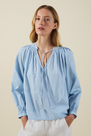 A woman wears the SYLVIE TOP by Velvet by Graham & Spencer—a light blue cotton poplin peasant blouse with a tie neckline and subtle floral embroidery—paired with white pants against a plain background.