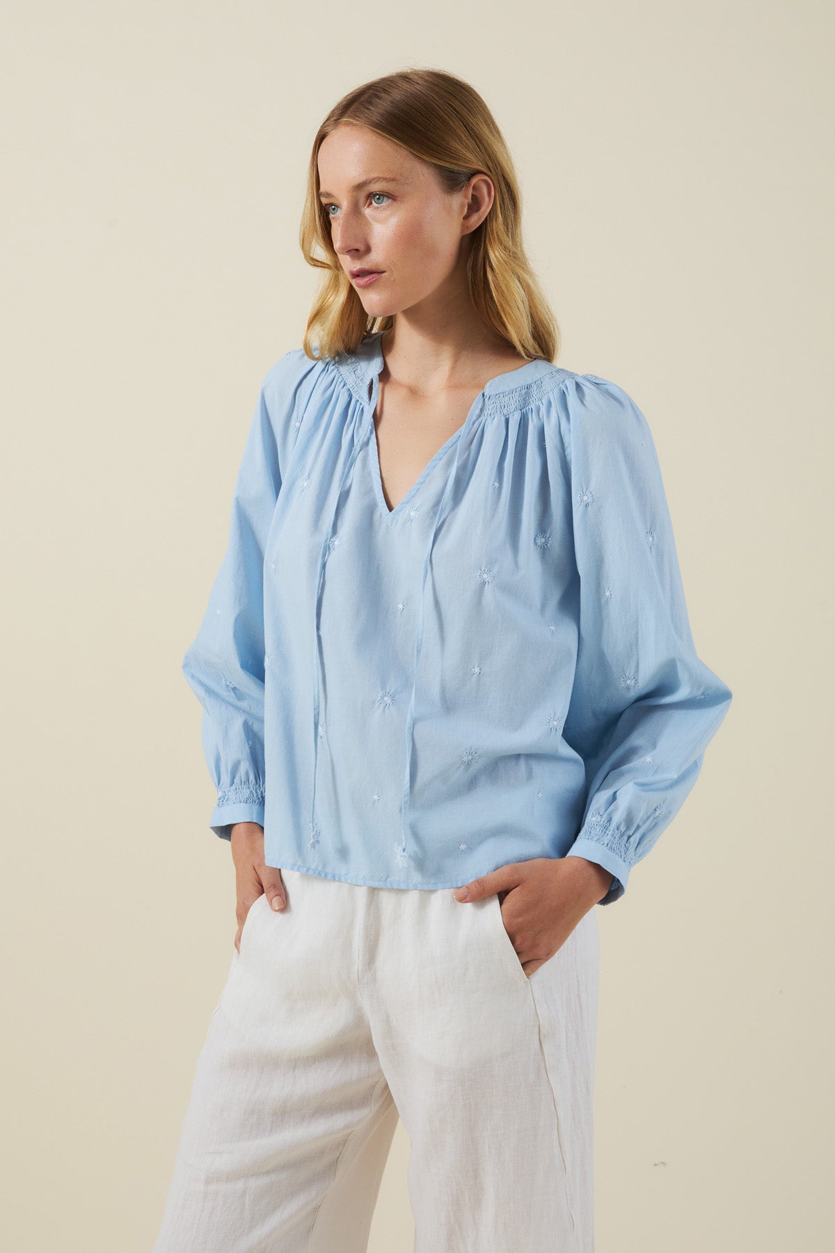 A person wears the SYLVIE TOP by Velvet by Graham & Spencer—a light blue cotton poplin peasant top with floral embroidery—paired with white pants, standing against a plain beige background.