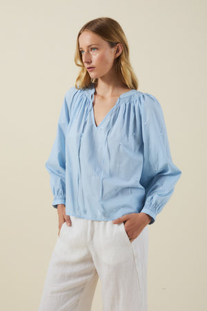A person wears the SYLVIE TOP by Velvet by Graham & Spencer—a light blue cotton poplin peasant top with floral embroidery—paired with white pants, standing against a plain beige background.