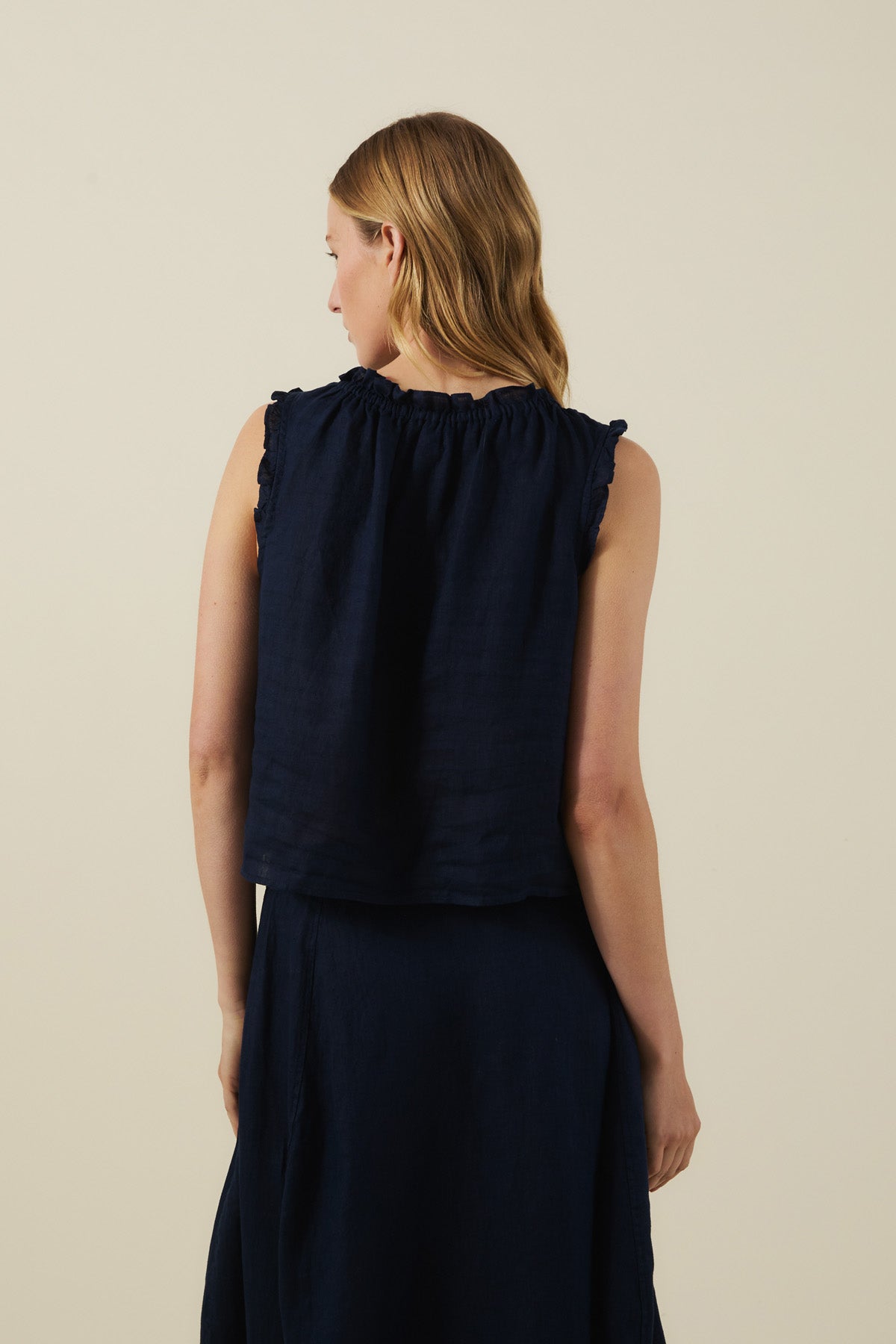 A woman with light brown hair is seen from the back against a plain light background, wearing the TANA LINEN TOP in dark navy by Velvet by Graham & Spencer with a matching skirt.-41527520985281