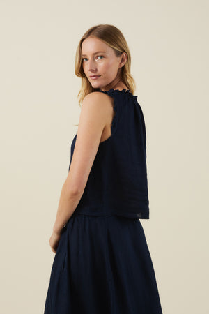 A woman wears the TANA LINEN TOP by Velvet by Graham & Spencer, featuring ruffled shoulders, paired with a matching navy skirt against a plain light background.