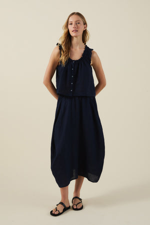 A woman stands against a plain background wearing the TANA LINEN TOP by Velvet by Graham & Spencer with a matching navy skirt and black sandals.