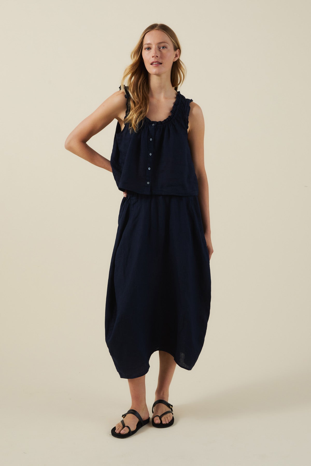 A woman stands against a plain background wearing the FAE LINEN SKIRT from Velvet by Graham & Spencer—a sleeveless navy top and matching A-line linen skirt—paired with black sandals.