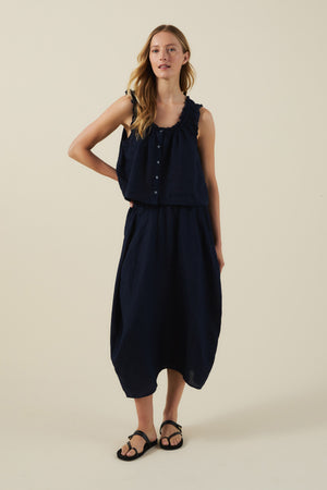 A woman stands against a plain background wearing the FAE LINEN SKIRT from Velvet by Graham & Spencer—a sleeveless navy top and matching A-line linen skirt—paired with black sandals.