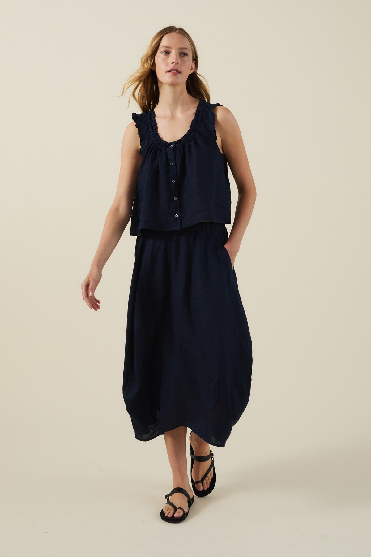 A woman stands against a plain beige background wearing Velvet by Graham & Spencer's FAE LINEN SKIRT with a sleeveless navy ruffle-detail top and black sandals.