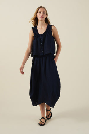 A woman stands against a plain beige background wearing Velvet by Graham & Spencer's FAE LINEN SKIRT with a sleeveless navy ruffle-detail top and black sandals.