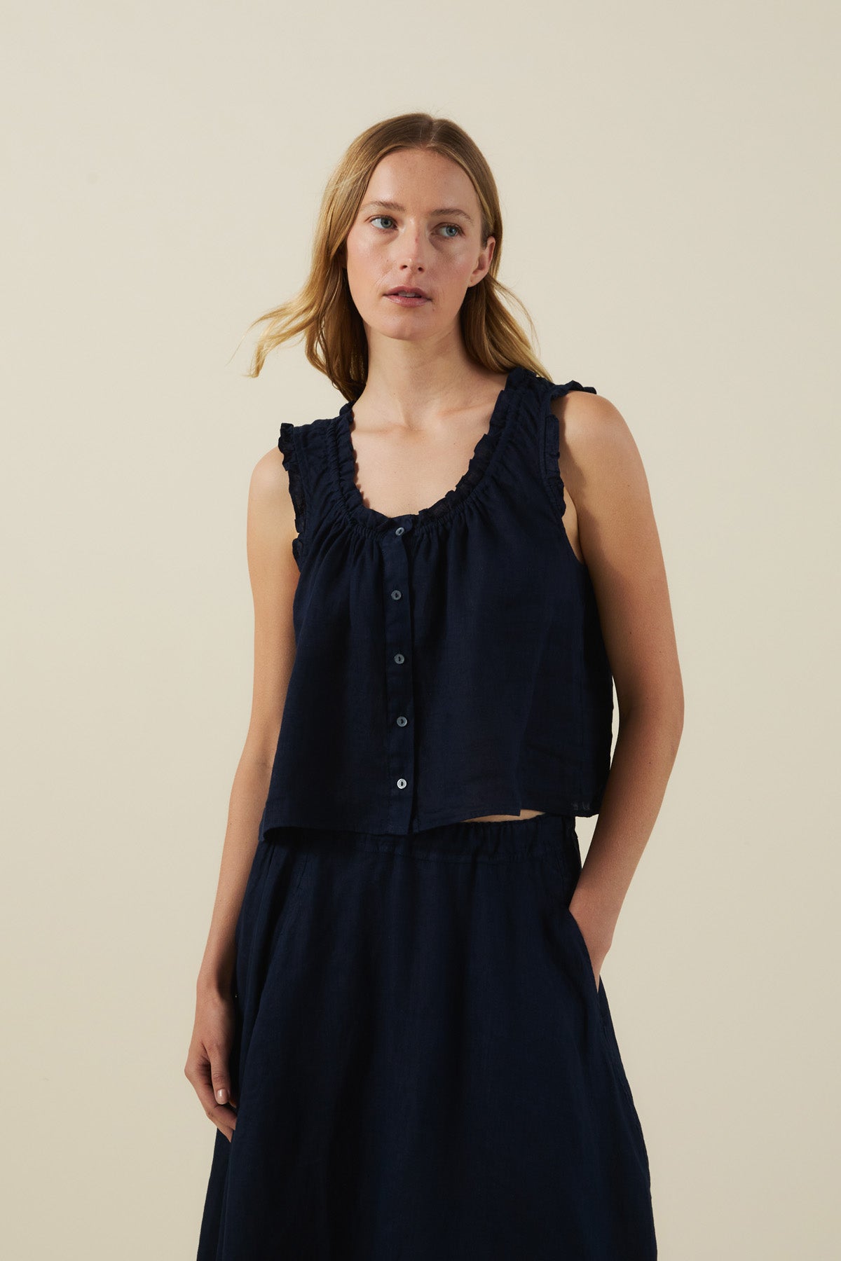 A woman wears the TANA LINEN TOP by Velvet by Graham & Spencer, a feminine navy button-front linen top with a ruffled neckline and matching skirt, standing against a plain light background.
