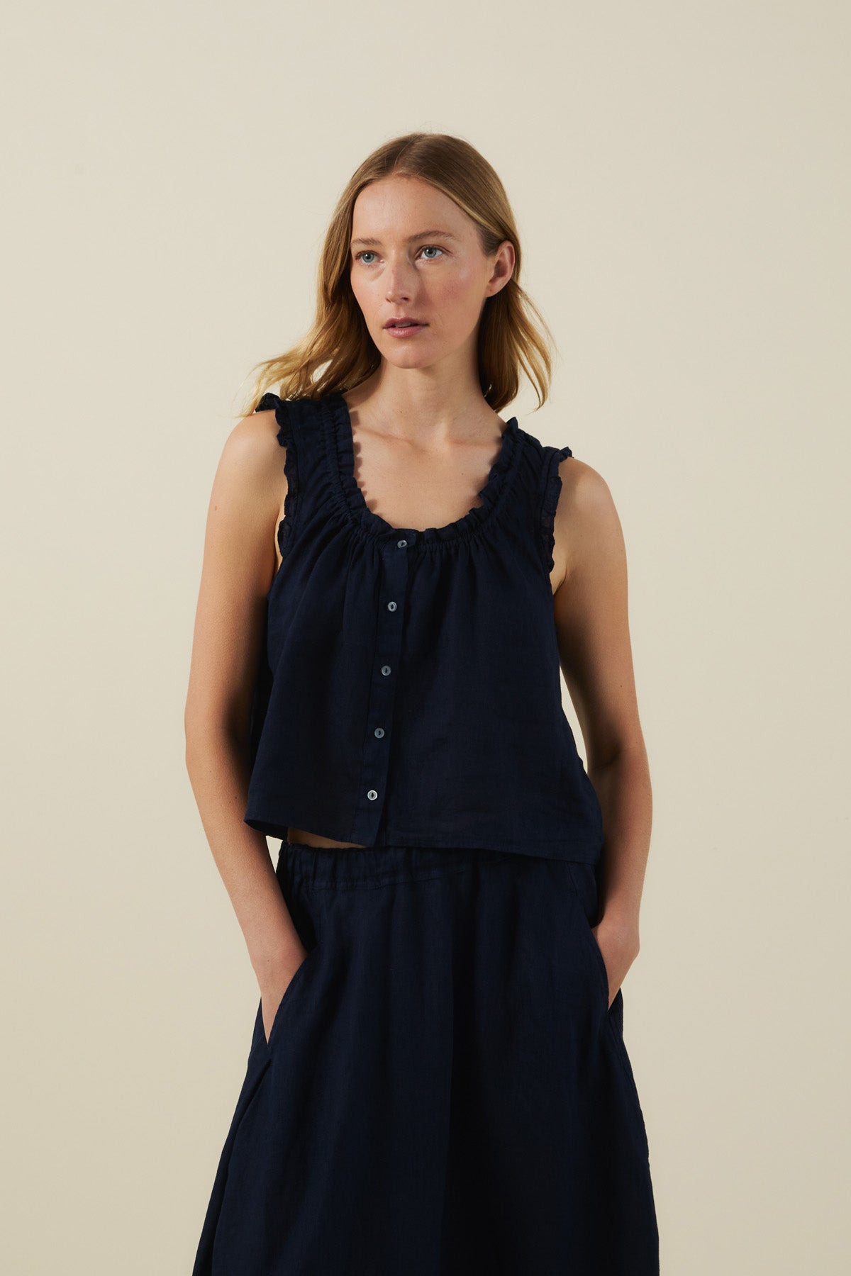 A woman wears the TANA LINEN TOP by Velvet by Graham & Spencer—a sleeveless, feminine linen button-front top paired with a matching skirt—standing with hands in pockets against a plain light background.-41527521247425
