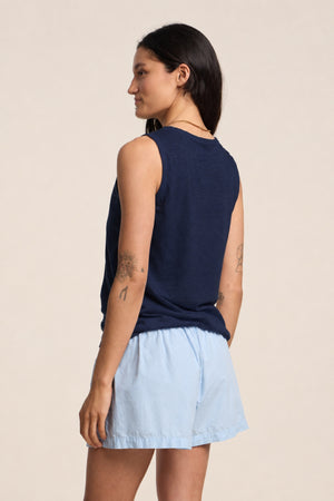 A person with long dark hair wears the TAURUS TANK TOP by Velvet by Graham & Spencer and light blue shorts, showing arm tattoos—a must-have for any relaxed summer look.