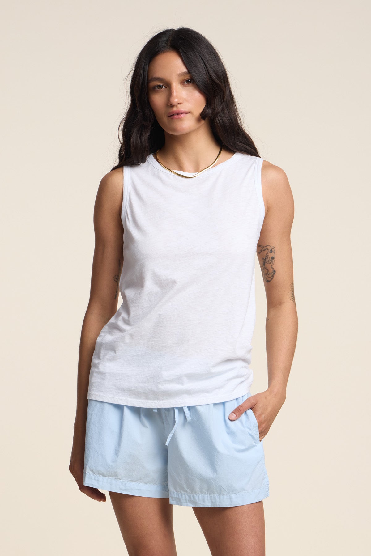 A woman with long dark hair wears the TAURUS TANK TOP by Velvet by Graham & Spencer—a white, crew-neck, sleeveless top in textured cotton slub—paired with light blue shorts, standing against a plain beige background with one hand in her pocket for an effortlessly stylish look.-41803096981697