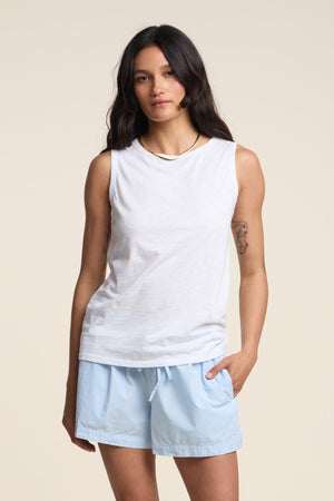 A woman with long dark hair wears the TAURUS TANK TOP by Velvet by Graham & Spencer—a white, crew-neck, sleeveless top in textured cotton slub—paired with light blue shorts, standing against a plain beige background with one hand in her pocket for an effortlessly stylish look.
